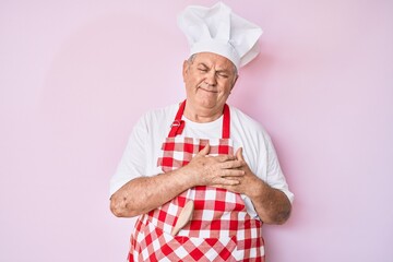 Senior grey-haired man wearing professional baker apron smiling with hands on chest with closed eyes and grateful gesture on face. health concept.