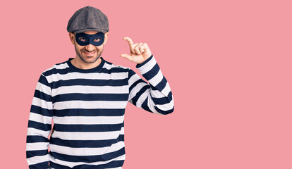 Young handsome man wearing burglar mask smiling and confident gesturing with hand doing small size sign with fingers looking and the camera. measure concept.