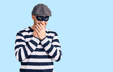 Young handsome man wearing burglar mask laughing and embarrassed giggle covering mouth with hands, gossip and scandal concept