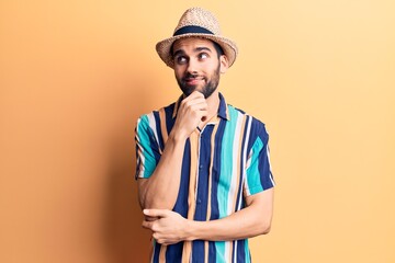 Young handsome man with beard wearing summer hat and shirt with hand on chin thinking about question, pensive expression. smiling with thoughtful face. doubt concept.