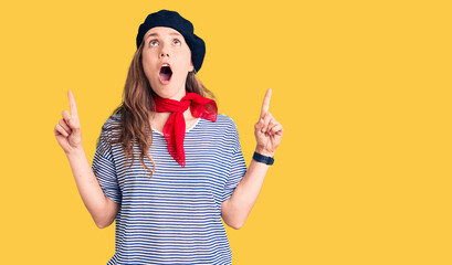 Young beautiful blonde woman wearing french beret and striped t-shirt amazed and surprised looking up and pointing with fingers and raised arms.