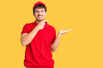 Young handsome man with curly hair wearing delivery uniform amazed and smiling to the camera while presenting with hand and pointing with finger.