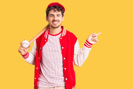 Young handsome man with curly hair playing baseball holding bat and ball smiling happy pointing with hand and finger to the side