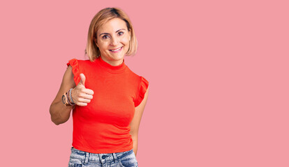 Young blonde woman wearing casual clothes doing happy thumbs up gesture with hand. approving expression looking at the camera showing success.