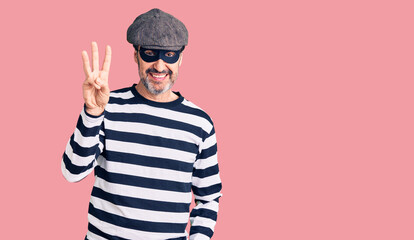 Middle age handsome man wearing burglar mask showing and pointing up with fingers number three while smiling confident and happy.