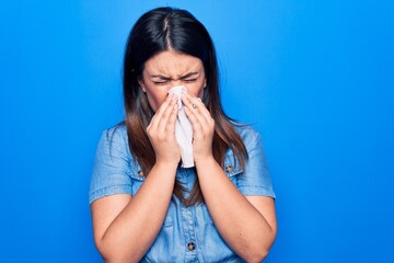 Young beautiful woman sick for flu. Using paper handkerchief on full nose of mucus standing over isolated blue background