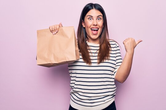 Young beautiful woman holding deliver paper bag with takeaway food over pink background pointing thumb up to the side smiling happy with open mouth