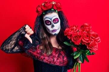 Young woman wearing day of the dead costume holding flowers with angry face, negative sign showing dislike with thumbs down, rejection concept