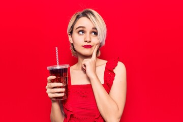 Young beautiful blonde woman drinking glass of cola beverage serious face thinking about question with hand on chin, thoughtful about confusing idea