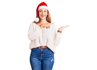 Beautiful young woman wearing christmas hat amazed and smiling to the camera while presenting with hand and pointing with finger.