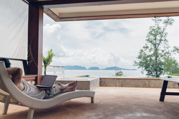 Asian man relax on the bench and working on his laptop with sea view.