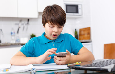 Naklejka premium Excited boy using smartphone while doing homework at kitchen