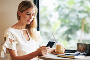 Young woman enjoying cold coffee beverage and reading test messages or articles on smartphone screen