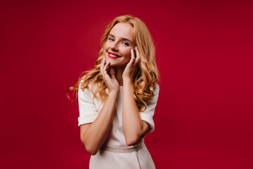 Pretty white woman in dress enjoying studio photoshoot. Blissful female model with blonde hair standing on red background.