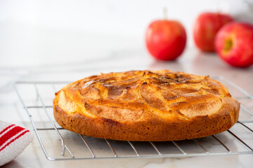 Homemade freshly baked Apple tea cake with cinnamon topping cooling on wire rack with red apples in background on marble kitchen counter top, selective focus