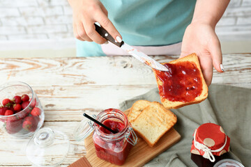 Woman making toasts with sweet strawberry jam in kitchen