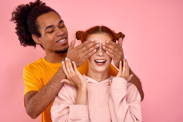 Handsome afro american guy wants to make a pleasant surprise for his beloved redhead girfriend of European appearance and closes her eyes with his hands. Concept of a happy relationship.