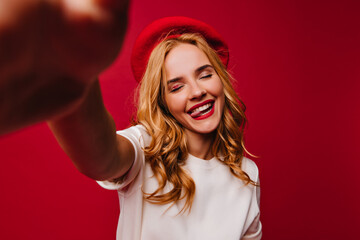 Naklejka premium Graceful french girl making selfie on red background with eyes closed. Indoor photo of debonair european woman posing with pleased face expression.
