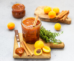 Apricot jam in a glass jar, fresh apricots, thyme and cinnamon on grey stone background.