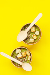Fresh broccoli cream soup with croutons and seeds in craft containers on yellow background. Soup to go, healthy food delivery.