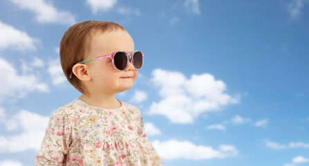 childhood, summer and people concept - happy little baby girl in sunglasses over blue sky and clouds background