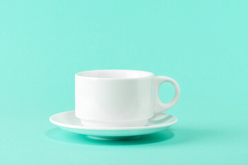 Empty porcelain coffee cup and a saucer over a mint green color background. White ceramic teacup. Modern stylish tableware. Copy space. Front view.
