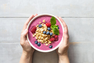 Female's hands holding smoothie bowl with granola and berries decorated with mint on grey wooden...