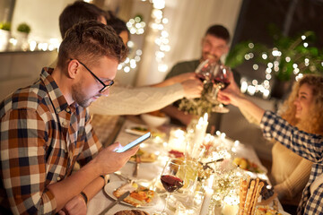 holidays, technology and people concept - young man in glasses with smartphone at christmas dinner party with friends at home