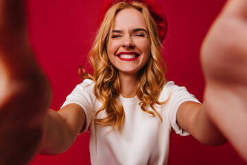 Laughing woman with wavy blonde hair making selfie on red background. Good-humoured french female model enjoying studio photoshoot.