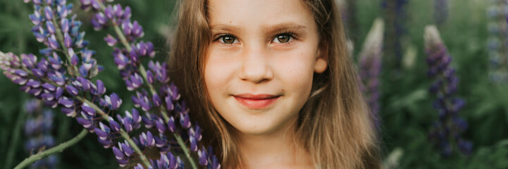 portrait of a cute little happy seven year old kid girl with bloom flowers lupines in a field in nature outdoor. banner