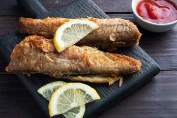 Fried fish hake Pollock with tomato sauce and slices of fresh lemon. Concept eating fatty fast food. Dark wooden background. Copy space.