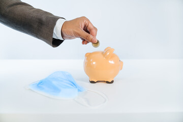 side view of a hand inserting a coin into a ceramic piggy bank with a white background and a face mask due to the covid19 coronavirus pandemic