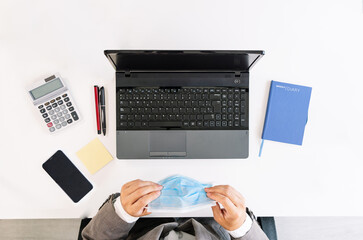 top view of a white office table with a laptop, calculator, cell phone, post it, pens, notebook and a business worker wearing a face mask