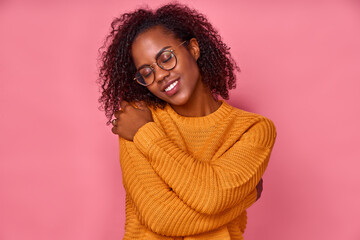 Close up portrait of a dark skinned woman with closed eyes and hugs herself, wears round glasses and orange sweater, posing in a romantic high spirit isolated over pink wall studio background.