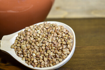 Buckwheat in a wooden spoon, next to a clay pot on a brown wooden background. The concept of healthy dietary food, vegetarianism, traditional Russian cuisine. Selective focus, place for text