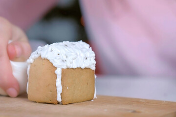 Woman is decorating gingerbread house with sugar sweet icing, hands closeup. Cooking, baking and decorating homemade gingerbread house for Christmas holidays. New Year family traditions.