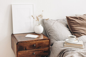 Portrait white frame mockup on retro wooden bedside table. Modern white ceramic vase, dry Lagurus ovatus grass. Cup of coffee and books in bed. Beige linen pillows in bedroom. Scandinavian interior.