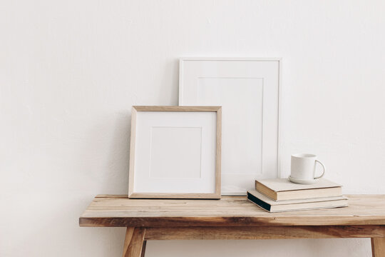 Wooden square and white portrait frame mockups on vintage bench, table. Cup of coffee on pile of books. White wall background. Scandinavian interior, neutral color palette. Artistic display concept.