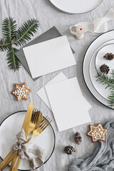 Fototapeta na wymiar Christmas table setting. Blank greeting cards mockup scene. Golden cutlery, green fir tree branches. Plates and gingerbread cookies, linen table cloth. Holiday background. Vertical flat lay, top view