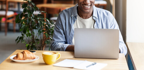 Cropped view of young black man using laptop for online work or studies at cafe, blank space. Panorama