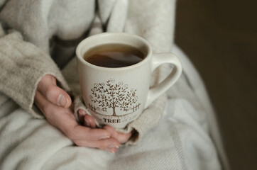Dark tea mug in female hands on beige plaid background. Warm autumn morning. Girl holds cup of dark tea 