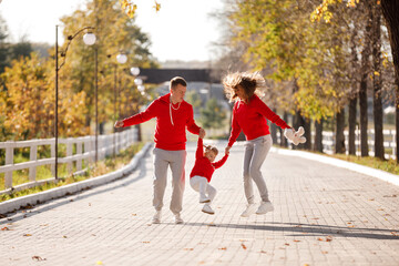 father and mother are holding little daughter by the hands and walking in the autumn park, happy family is having fun outdoors. father's, mother's and baby's day