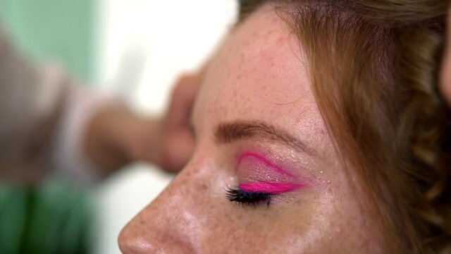 Unrecognizable makeup artist work in beauty studio. Woman applying tone using brush. Artist make a makeup for redhead model with freckles. Close up of a pretty girl with pink eye shadows