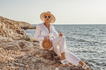 Outdoor summer fashion portrait of elegant woman wearing white linen suit, straw hat, sunglasses,...