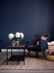 Bright interior of the living room. Blue wall, gray chair and coffee table. Natural decor woven basket. White peonies in a glass vase