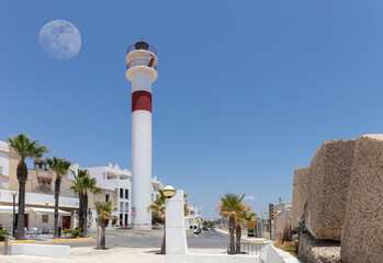 The red and white Rota lighthouse with the moon in the sky. There are white houses, restaurants and some palm trees. On the right are some concrete blocks from the pier.