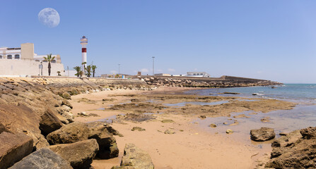 The red and white lighthouse of Rota stands behind a bay. In the foreground the beach with sand and...