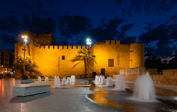 In the evening in the Spanish palm city of Elche. The castle is in the blue hour with water fountains in the foreground. There are clouds in the sky with some red sunset on the horizon.