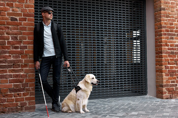 portrait of blind man leaned on red brick building, wearing dark eyeglasses, holding guide dog on...