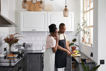 attractive african lady hugs her husband while he is preparing food in the kitchen. married couple in love, woman admires her man